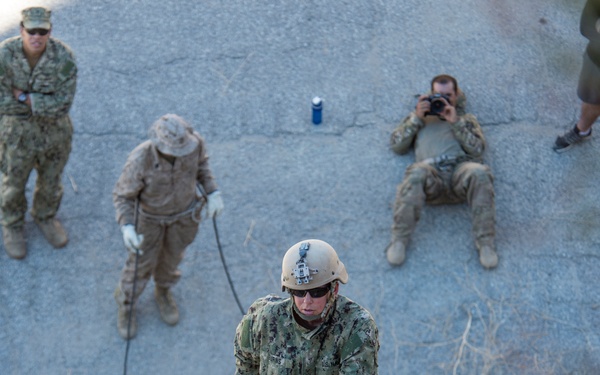 A military service member in U.S. Navy NWU Type III camouflage uniform is rappelling down a rocky or concrete incline during a training exercise. Several observers in various military uniforms watch from above, including one individual photographing the activity. The scene appears to be a controlled outdoor training environment.