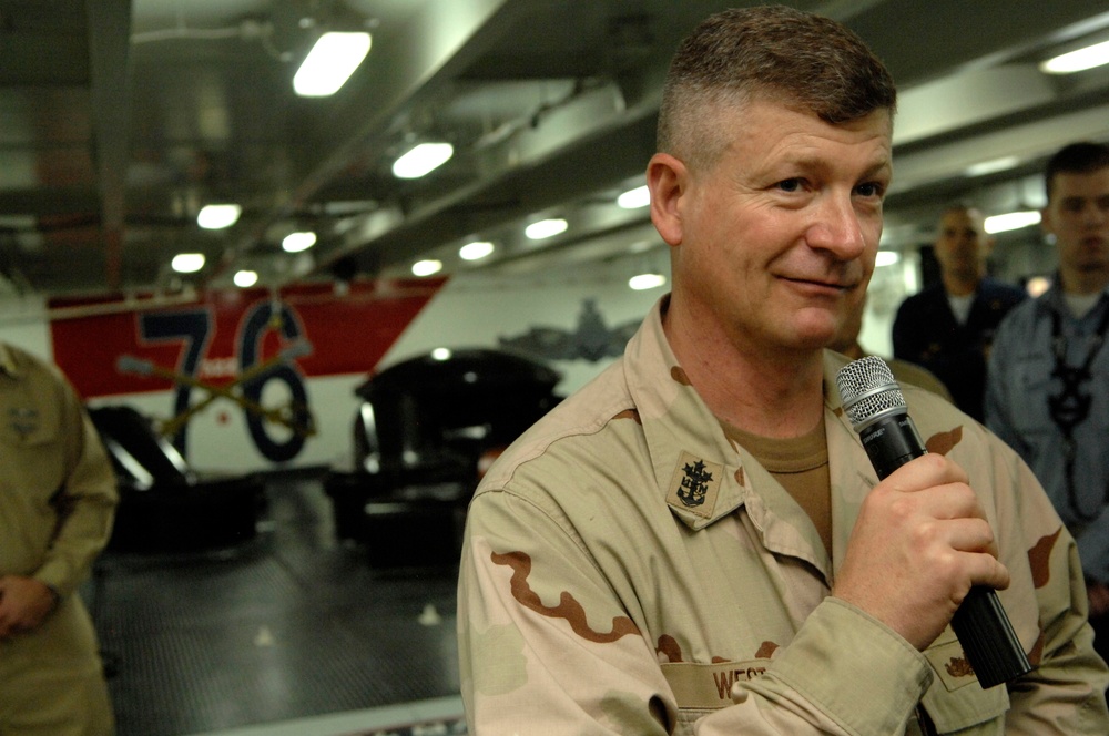 A military personnel member in desert camouflage (DCU) uniform is speaking into a handheld microphone, likely addressing an audience. The setting appears to be the hangar bay of a naval vessel, indicated by the number '76' visible on what appears to be an aircraft or vehicle in the background. Other uniformed personnel are visible in the background.
