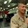 A military personnel member in desert camouflage (DCU) uniform is speaking into a handheld microphone, likely addressing an audience. The setting appears to be the hangar bay of a naval vessel, indicated by the number '76' visible on what appears to be an aircraft or vehicle in the background. Other uniformed personnel are visible in the background.