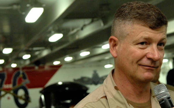A military personnel member in desert camouflage (DCU) uniform is speaking into a handheld microphone, likely addressing an audience. The setting appears to be the hangar bay of a naval vessel, indicated by the number '76' visible on what appears to be an aircraft or vehicle in the background. Other uniformed personnel are visible in the background.