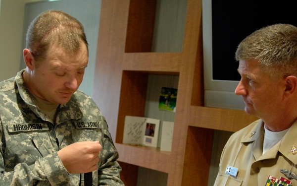 Two U.S. military personnel are engaged in a conversation indoors. The soldier on the left, identified by his name tape as 'HROMYSIN' of the U.S. Army, is holding a white cane and wearing ACU (Army Combat Uniform) with a 2nd Infantry Division patch. The officer on the right is wearing a U.S. Navy khaki service uniform with multiple ribbons and insignia.