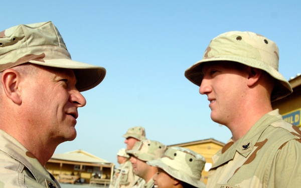 A military ceremony is taking place outdoors in what appears to be a desert environment. A senior officer in desert camouflage (DCU pattern) is pinning a medal or award onto the uniform of a US Navy sailor wearing a boonie hat. Several other uniformed personnel are visible in the background observing the ceremony.