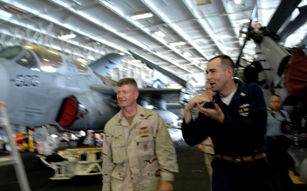 Two military personnel are walking through what appears to be the hangar bay of a US Navy aircraft carrier. One individual is wearing desert camouflage (DCU) uniform while the other is in Navy service dress blue uniform, suggesting a tour or briefing is taking place. Military aircraft, including one with the number '506' visible, are stored in the background.