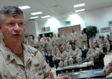 A U.S. Navy servicemember named 'WEST' is addressing a large group of military personnel in what appears to be a common room or briefing area. The speaker is in the foreground wearing desert camouflage (DCU) uniform with visible rank insignia and Navy tape. A crowd of approximately 30-40 service members in similar desert uniforms are seated and standing in the background, attentively listening.