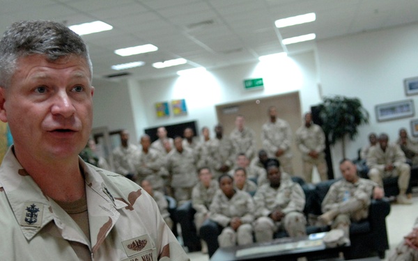A U.S. Navy servicemember named 'WEST' is addressing a large group of military personnel in what appears to be a common room or briefing area. The speaker is in the foreground wearing desert camouflage (DCU) uniform with visible rank insignia and Navy tape. A crowd of approximately 30-40 service members in similar desert uniforms are seated and standing in the background, attentively listening.