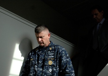 A U.S. Navy servicemember in digital camouflage uniform descends a staircase, with a civilian man in a dark suit visible in the shadowed background. The lighting is dramatic, with strong contrast between light and shadow. The Navy personnel appears to be walking downstairs while holding the handrail.