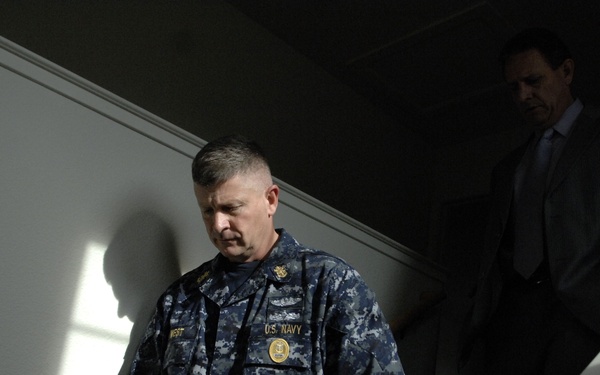 A U.S. Navy servicemember in digital camouflage uniform descends a staircase, with a civilian man in a dark suit visible in the shadowed background. The lighting is dramatic, with strong contrast between light and shadow. The Navy personnel appears to be walking downstairs while holding the handrail.
