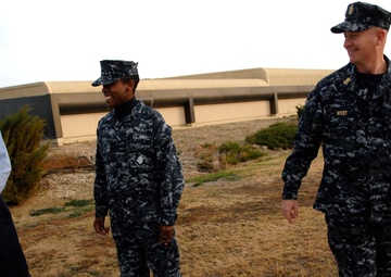 Three U.S. Navy personnel are standing outdoors near a large building, appearing to be engaged in a casual conversation. Two individuals are wearing the Navy Working Uniform (NWU) digital camouflage pattern, while one person on the left is wearing a light blue Navy shirt and a black 'NAVY' cap. The person on the right has a name tape reading 'WEST' and displays officer insignia and medals.