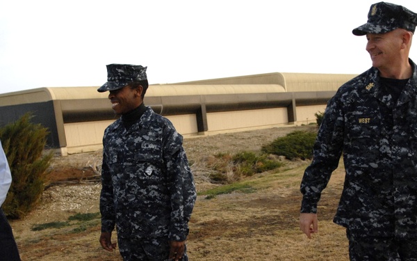 Three U.S. Navy personnel are standing outdoors near a large building, appearing to be engaged in a casual conversation. Two individuals are wearing the Navy Working Uniform (NWU) digital camouflage pattern, while one person on the left is wearing a light blue Navy shirt and a black 'NAVY' cap. The person on the right has a name tape reading 'WEST' and displays officer insignia and medals.