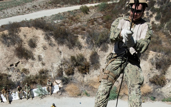 A military service member in camouflage uniform is rappelling down a rocky cliff face using a rope and harness system. Below on a concrete or paved surface, multiple other personnel are observing or waiting their turn, suggesting this is a training exercise. The arid, scrub-covered terrain and dry hillside indicate a southwestern United States training environment.