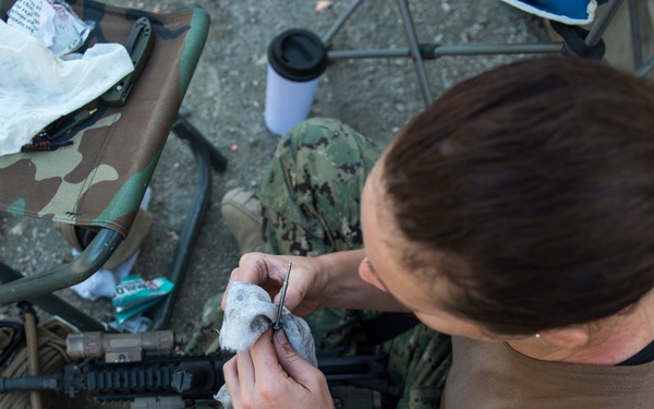 A military service member in Navy Working Uniform (NWU) camouflage is performing weapon maintenance on what appears to be an M4/M16-style rifle. The individual is seated on the ground and using a cleaning rod and cloth patch to clean the barrel or components of the firearm. Various military gear, a tactical bag, folding chairs, and cleaning supplies are visible in the surrounding area.