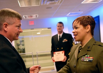 A formal military ceremony is taking place in what appears to be a 'Hall of Heroes' or similar official venue. A civilian man in a dark suit is shaking hands with a female U.S. Marine in dress green uniform while passing what appears to be a coin or small item. A Navy officer stands in the background holding a red-bordered certificate or award document.