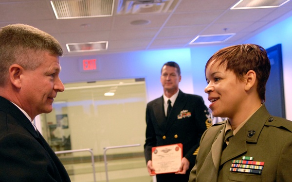 A formal military ceremony is taking place in what appears to be a 'Hall of Heroes' or similar official venue. A civilian man in a dark suit is shaking hands with a female U.S. Marine in dress green uniform while passing what appears to be a coin or small item. A Navy officer stands in the background holding a red-bordered certificate or award document.