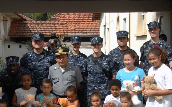 A group of U.S. Navy personnel in digital camouflage uniforms pose with local children, adults, and an officer in a gray dress uniform. The children are holding stuffed animals, suggesting a community outreach or humanitarian assistance event. The setting appears to be an outdoor area near a residential or institutional building in what appears to be a Latin American or Caribbean location.