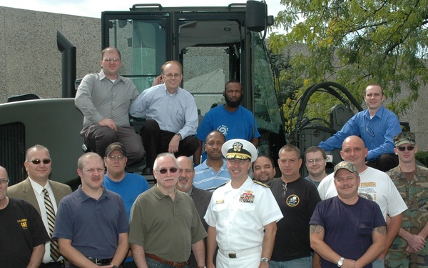 A group of approximately 18 men pose for a formal group photograph in a parking lot in front of a large military or industrial vehicle. The group includes a U.S. Navy officer in dress whites at the center, civilians in business and casual attire, and at least two individuals in military camouflage uniforms. Three men are seated on top of the large green vehicle while the rest stand in front of it.