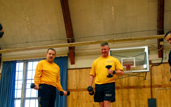 A group of U.S. Navy personnel are conducting a physical training session inside a gymnasium. They are wearing matching yellow 'NAVY' shirts and dark navy shorts while exercising with dumbbells. The low-angle shot captures multiple participants mid-exercise with additional dumbbells visible on the gym floor in the foreground.