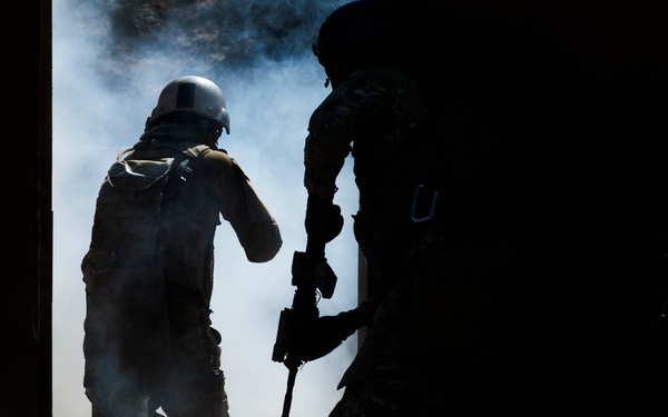 The image depicts two armed personnel in tactical/military gear silhouetted against a smoke-filled doorway or breach point. One figure is partially illuminated, wearing a helmet and carrying equipment on their back, while a second figure in the foreground holds a rifle. The scene is dramatically backlit with dust and smoke creating a hazy atmosphere.