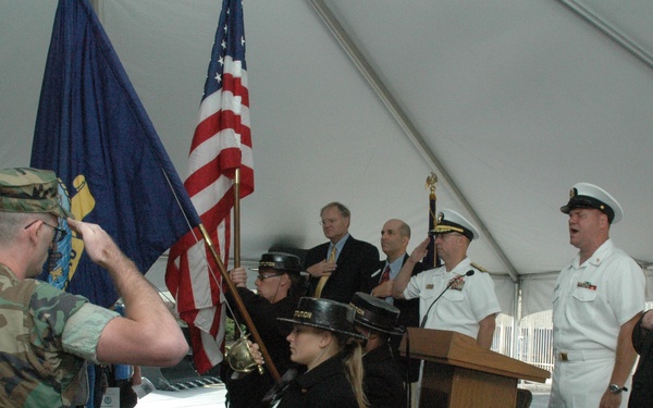 A formal military or civic ceremony is taking place under a large white tent. The scene includes a color guard presenting flags, military personnel in dress whites, a soldier in camouflage saluting, and civilians standing at attention near a wooden podium. The event appears to be an official commissioning, dedication, or patriotic ceremony.