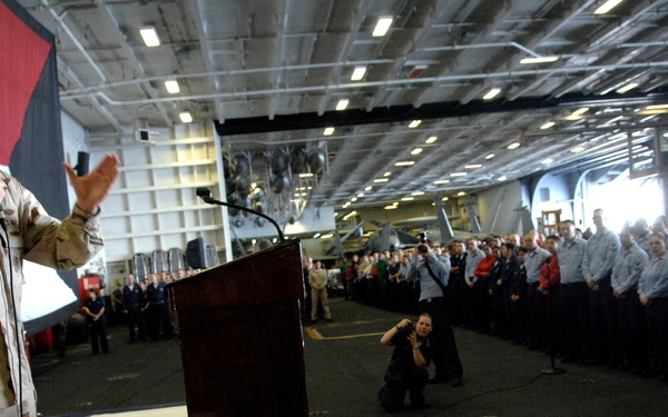 A military officer in desert camouflage (DCU) uniform is addressing a large formation of naval personnel in what appears to be the hangar bay of an aircraft carrier. The speaker stands at a wooden podium holding a microphone, with rows of sailors in blue uniforms standing at attention. A photographer is crouched nearby capturing the event, and military aircraft are visible in the background.