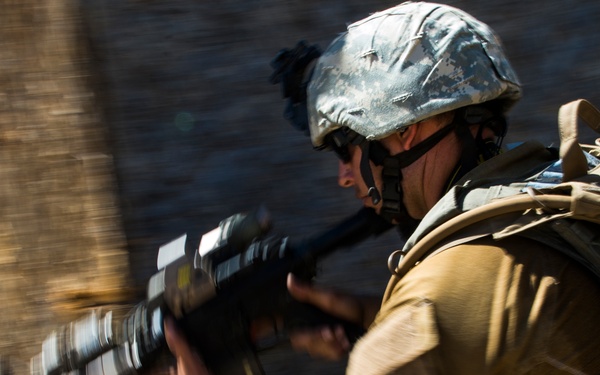 A soldier in full combat gear is captured in motion, moving along what appears to be a wall or building exterior. The image features intentional motion blur, suggesting rapid movement during what appears to be a tactical operation or training exercise. The soldier is equipped with a helmet, tactical vest, backpack, and is holding a rifle in a ready position.