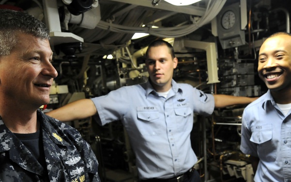 Three U.S. Navy personnel are gathered in what appears to be the interior of a naval vessel, likely a submarine or ship's engineering space. The personnel are engaged in a casual conversation, with two wearing light blue Navy working uniforms and one in Navy Working Uniform (NWU) digital camouflage. The atmosphere appears relaxed and positive, with at least one sailor visibly laughing.
