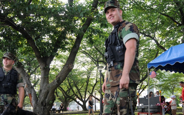Two military or law enforcement personnel in camouflage uniforms and tactical vests are handling K-9 dogs in a park setting. One handler stands with a black German Shepherd lying on the grass in the foreground, while a second handler stands with a tan German Shepherd in the background. The scene appears to be a public outdoor event or demonstration, with civilians and a blue canopy tent visible to the right.
