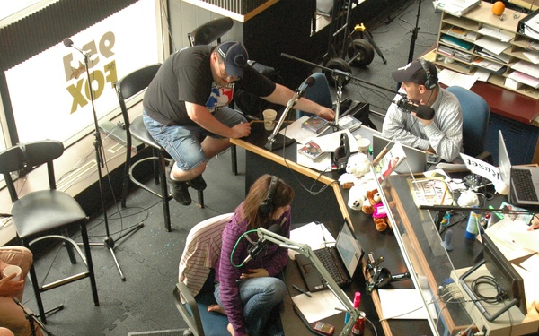 An overhead view of a busy radio station studio showing multiple people working at desks equipped with microphones, headphones, and laptops. The studio appears to be for 'Free Fox' based on reversed text visible on the window, with a station identifier showing '1180 WHAM' also visible. Several on-air personalities and staff are engaged in what appears to be a live broadcast session.
