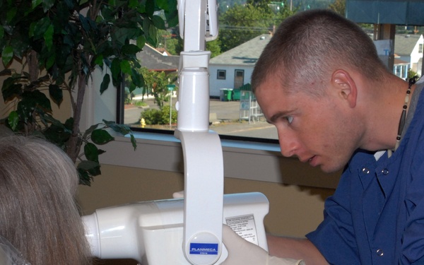 A dental professional in blue scrubs and white latex gloves is positioning a dental X-ray machine (branded 'Planmeca') near a seated patient's head. The patient, an older woman with gray-brown hair, is seated in a dental chair covered with a protective plastic drape and wearing a lead apron bib. The setting appears to be a dental office with a window overlooking a street.