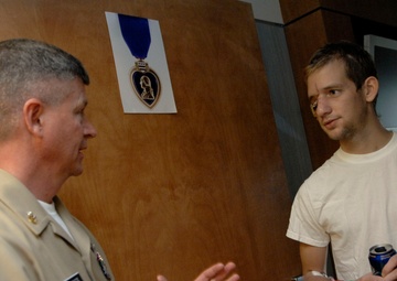 A military officer in tan/khaki uniform is speaking with a young civilian man in a white t-shirt, who is holding a beverage can. They appear to be in a medical or administrative facility, as evidenced by the hospital-style wristbands on the civilian's wrist. A printed image of the Purple Heart medal is visible on the wooden door behind them.