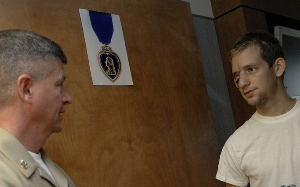 A military officer in tan/khaki uniform is speaking with a young civilian man in a white t-shirt, who is holding a beverage can. They appear to be in a medical or administrative facility, as evidenced by the hospital-style wristbands on the civilian's wrist. A printed image of the Purple Heart medal is visible on the wooden door behind them.