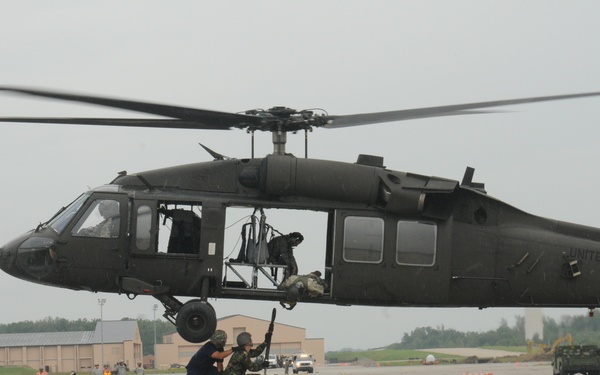 A United States Army UH-60 Black Hawk helicopter hovers low over a military airfield tarmac. Ground personnel are positioned near a wheeled cart or trailer directly beneath the aircraft, appearing to conduct a loading or equipment transfer operation. Additional military personnel and vehicles are visible in the background along the flight line.