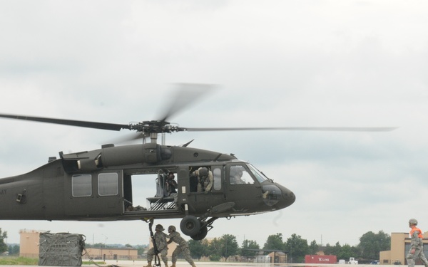 A United States Army UH-60 Black Hawk helicopter hovers low over a military airfield tarmac while ground personnel interact with it. Several soldiers in military uniforms are visible on the ground, with one appearing to attach or detach a sling load beneath the aircraft. The scene appears to depict a sling load or external lift training exercise at a military installation.