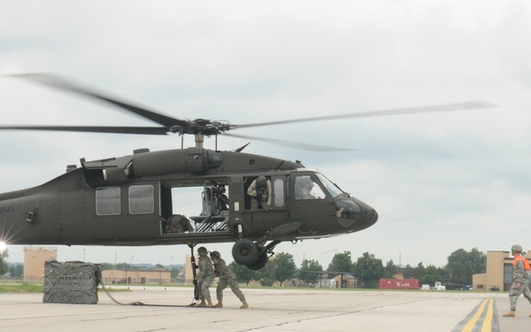 A United States Army UH-60 Black Hawk helicopter is on a military airfield tarmac with rotor blades spinning. Ground personnel in military uniforms are actively working around the aircraft, appearing to conduct a sling load or cargo hook operation. The scene takes place at what appears to be a military installation with hangars and support vehicles visible in the background.