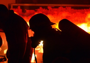 The image shows multiple firefighters silhouetted against an intense fire burning behind a mesh or grid structure. The scene is dominated by deep orange and red tones from the flames, creating dramatic backlighting that renders the figures almost entirely in shadow. At least one firefighter is visibly equipped with a breathing apparatus (SCBA tank) and appears to be in a crouched position.
