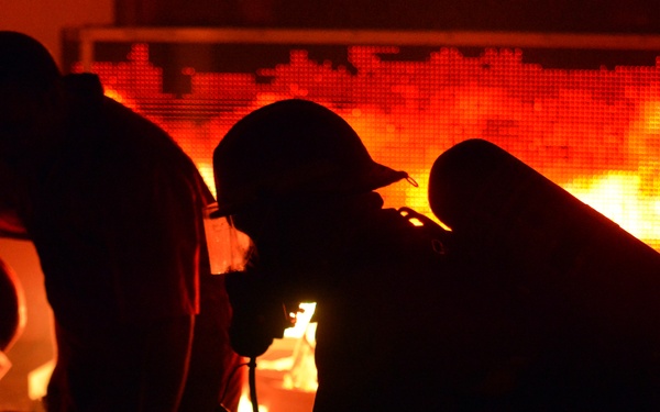 The image shows multiple firefighters silhouetted against an intense fire burning behind a mesh or grid structure. The scene is dominated by deep orange and red tones from the flames, creating dramatic backlighting that renders the figures almost entirely in shadow. At least one firefighter is visibly equipped with a breathing apparatus (SCBA tank) and appears to be in a crouched position.