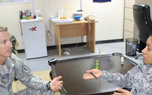 Two U.S. Army soldiers in ACU (Army Combat Uniform) camouflage are engaged in what appears to be a counseling or administrative meeting in an office setting. The female soldier on the right, who appears to hold the rank of Captain (visible rank insignia), is holding papers and a pen while the male soldier on the left gestures as he speaks. The setting suggests a formal but informal consultation, possibly a counseling session or briefing.