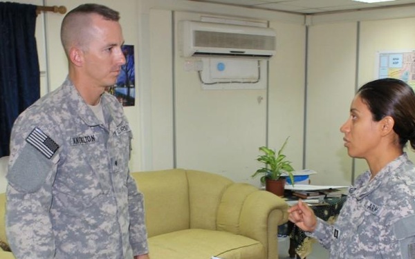 Two U.S. Army soldiers in ACU (Army Combat Uniform) are engaged in what appears to be a briefing or discussion inside a military office or common area. The female soldier on the right is holding documents and appears to be presenting or reviewing information with the male soldier on the left. The setting suggests a forward operating base or administrative military facility.
