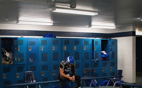 A lone football player sits on a bench in a locker room, head bowed and still wearing his blue helmet and uniform. The room is otherwise empty, with clothing and gear scattered across the floor and benches, suggesting the rest of the team has already left. The atmosphere conveys solitude and emotional weight, likely following the conclusion of a game.