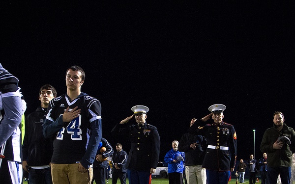 A nighttime ceremony on a football field showing a group of people standing at attention, likely during a national anthem. The group includes football players in blue uniforms, two military personnel in dress uniforms saluting, and civilian spectators. One player on the left is on crutches and has his hand over his heart.