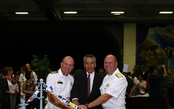 Three men — two in white U.S. Navy officer uniforms and one in a civilian suit — pose while cutting a large elaborately decorated cake shaped like an aircraft carrier bearing the hull number '73'. The event appears to be a formal military celebration or commissioning ceremony held in a large indoor venue. A crowd of military personnel and civilians is visible in the background.