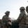 Two or more U.S. Army soldiers in Operational Camouflage Pattern (OCP) uniforms are engaged in what appears to be a training exercise or briefing at an outdoor range or obstacle course. The soldier on the left is wearing a combat helmet with goggles and a communications headset, and displays a U.S. flag patch and a unit patch on the left sleeve. Stacked vehicle tires are visible in the background, commonly used as range barriers or training obstacles.