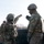 Two military personnel in full combat gear are engaged in a discussion or briefing at what appears to be a training area or obstacle course. Both are wearing MultiCam/OCP camouflage uniforms, tactical vests, and helmets. One soldier is gesturing with a raised fist while the other listens, with stacked large vehicle tires visible in the background.