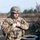 A U.S. Army soldier in full combat gear is standing at what appears to be a training range or field exercise area. The soldier is wearing MultiCam (OCP) camouflage uniform with a tactical helmet, body armor, and magazine pouches, and appears to be adjusting or checking equipment on their chest. The background features stacked tires used as range barriers, sparse trees, and an overcast sky suggesting a temperate climate.