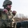 A U.S. Army soldier in full combat gear, identified by the name tape 'MANALTO' on their plate carrier, appears to be demonstrating or explaining a hand-to-hand combat or tactical movement technique to another soldier. The soldier is wearing MultiCam (OCP) camouflage uniform, a combat helmet with accessories, protective eyewear, and a neck gaiter. The setting appears to be an outdoor training environment with bare trees and earthen berms visible in the background.