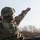 A U.S. Army soldier identified by the name tape 'PIANALTO' is in a field training environment, appearing to throw or gesture with an extended arm while holding an object in the other hand. The soldier is wearing MultiCam (OCP) camouflage uniform, body armor, a combat helmet with accessories, protective eyewear, and a neck gaiter. A second soldier in similar gear is partially visible on the right side of the frame.