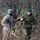 Two military personnel in a wooded outdoor setting appear to be greeting each other with a fist bump or handshake. One soldier wears US Army MultiCam (OCP) uniform with a visible American flag patch, while the other wears a darker woodland camouflage pattern with a green beret and what appears to be an Estonian flag patch. The setting suggests a joint military exercise or training event during late autumn.