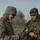 Two soldiers, one wearing a US Army uniform with an American flag patch and another wearing an Estonian Defense Forces uniform with an Estonian flag patch, are examining a small object together in an outdoor field setting. A third soldier in camouflage with a balaclava stands in the background holding a rifle. The scene appears to depict a joint military exercise or training event between US and Estonian forces.