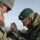 Two military personnel from different nations are engaged in a close interaction, with one appearing to pin or attach something to the other's uniform. The soldier on the left wears US Army MultiCam (OCP) uniform with a 1st Cavalry Division patch and US flag patch. The soldier on the right wears Estonian Defence Forces digital camouflage with an 'ESTONIA' shoulder tab and national flag patch, and a dark green beret.