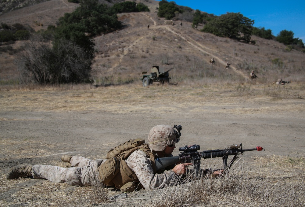 A U.S. Marine in MARPAT camouflage is in a prone firing position with an M16/M4-style rifle equipped with a bipod and optic scope. Additional personnel and a military vehicle are visible in the background, with other figures ascending a hillside trail. The setting appears to be a dry, arid training range consistent with a California military installation.