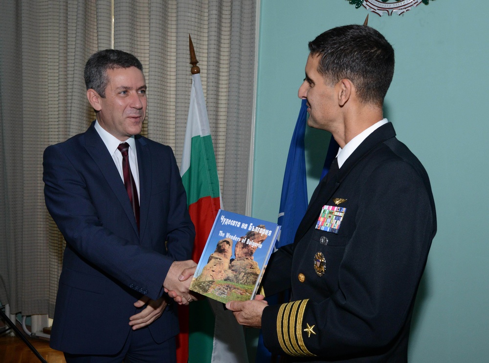 Two men are engaged in a formal exchange, with a civilian in a dark suit presenting a book titled 'The Wonders of Bulgaria' (Чудесата на България) to a military officer in a dark naval uniform adorned with medals and rank insignia. The setting appears to be an official meeting room with a Bulgarian national flag and what appears to be a NATO flag visible in the background. The interaction suggests a diplomatic or ceremonial gifting occasion.